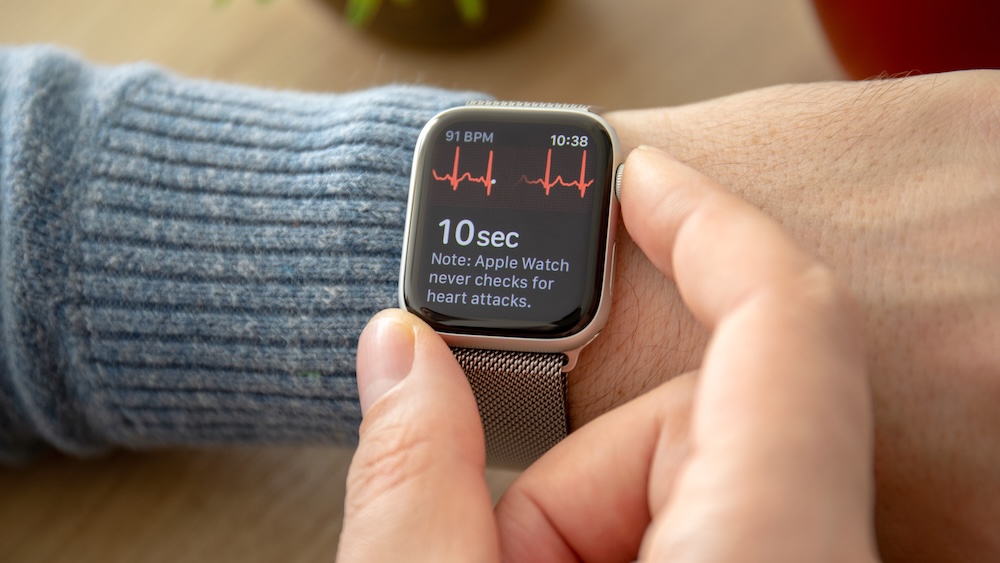 A hand holding an Apple Watch with two fingers during an ECG recording, index finger on the Digital Crown, thumb steadying the watch body
